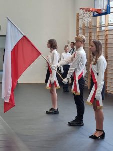 Zdjęcie ustępującego pocztu flagowego. Uczniowie mają przełożone biało-czerwone szarfy a uczeń stojący po środku trzyma flagę Polski.