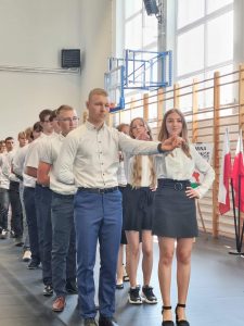 Uczniowie klasy ósmej stoją w parach podczas tańczenia poloneza. Chłopcy mają prawą rękę schowaną za plecami a dziewczynki opartą na biorze. Lewe ręce uczniowie mają złączone i wyciągnięte do przodu.
