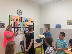 Na szkolnej stołówce wychowawczyni klasy czwartej trzyma w dłoniach przed sobą tort z zapalonymi świeczkami. Na przeciwko stoi solenizantka oraz jej koledzy.