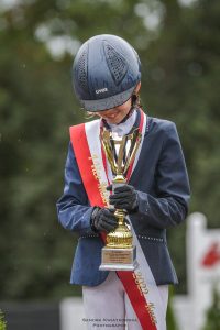 Uczennica Maja Iwińska na pochylonej glowie ma kask jeździecki a w dłoniach trzyma puchar.