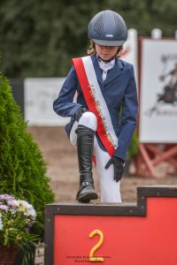 Uczennica Maja Iwińska wchodzi na podium z numerem 2. Jedna jej noga stoi na podwyższeniu a na drugiej wyprostowanej trzyma dłoń w rękawiczce.