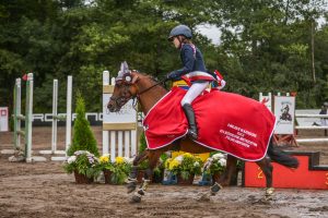 Uczennica Maja Iwińska siedzi na koniu w galopie. W tle widac ustawione przeszkody. Koń jest ubrany w czerwoną pelerynę z białym napisem w dolnym rogu: 'II MIEJSCE W KATEGORII KUCE XXV JEDZIECKIE MISTRZOSTWA POLSKI AMATORÓW".