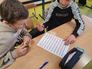 Dwaj uczniowie siedzą przy wspólnym stoliku trzymając w dłoniach kolorowe pisaki. przed nimi leży kartka z ustawionymi w rzedy kropkami.