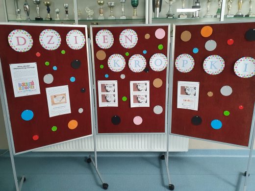 Zdjęcie gazetki przygotowanej z okazji dnia kropki. Na mobilnej tablicy umieszczono napis DZIEŃ KROPKI - każda litera została wydrukowana w osobnym kółeczku. Pod spodem umieszczono kolorowe kropki oraz karty z informacjami na temat dnia kropki.