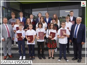 Stypendyści Gminy Łysomice pozują dozdjęcia wraz z wójtem Gminy Łysomicem i przewodniczącym Rady Gminy. Panowie stoją osobno a pomiędzy nimi ustawieni są uczniowie trzymający w dłoniach teczki z otrzymanymi stypendiami. Zdjęcie wykonane przed budynkiem urzędu Gminy Łysomice, dodatkowe ozdobione białą ramką oraz podpisem GMINA ŁYSOMICE w lewym rogu i herbem gminy w prawym górnym rogu.