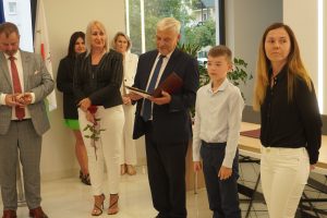 W rzędzie obok siebie stoją przedstawiciele gminy Łysomice oraz uczeń Tomasz Masny wraz z mamą. Obok ucznia po lewej stronie stoi wójt trzymający dla ucznia dyplom.