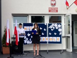 Przy głównym wejściu do szkoły stoi dwoje uczniów klasy ósmej prowadzących inagurację roku szkolnego 2022/2023. Za nimi na niebieskiej tablicy ozdobionej białymi kwiatami i motylami umieszczono napis; WITAJ W SZKOLE. Obok uczniów stoi statyw z flagami Polski.