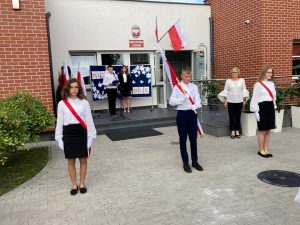 Na tle głównego wejści do szkoły stoi poczet flagowy. Za nimi w tle widać dwoje uczniów klasy ósmej prowadzących inagurację roku szkolnego 2022/2023.