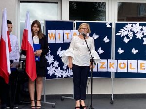 Pani dyrektor stoi za mikrofonem ustawionym na statywie. Za nią widać tablice z napisem: WITAJ W SZKOLE oraz dwoje uczniów klasy ósmej prowadzących inaugurację roku szkolnego 2022/2023.