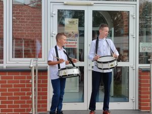 Dwóch uczniów klasy ósmej stoi przed bocznymi drzwiami do szkoły. Chłopcy maja zawieszone na szyi białe bębenki a w rękach pałeczki. Obaj mają wzrok skierowany w prawą stronę.