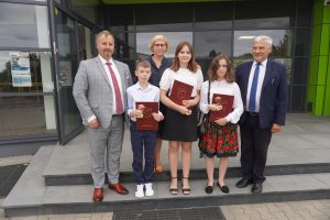 Troje stypendystów pozuje do zdjęcia wraz z wójtem Gminy Łysomice, przewodniczącym Rady Rodziców oraz dyrektorką Szkoły Podstawowej w Ostaszewie. Uczniowie stoją w jednym rzędzie w towarzystie przedstawicieli gminy a za nimi stoi Pani Dyrektor. Zdjęcie wykonane na stołach przed budynkiem Urzędu Gminy.