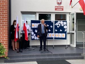 Wójt Gminy Łysomice podczas przemowy na inauguracji roku szkolnego 2022/2023. Wójt stoi za statywem z mikrofonem. Za nim ustawiono granatową tablicę ozdobioną bialymi kwiatami i motylami z napisem : WITAJ W SZKOLE. Zdjęcie wykonane przed głównym weejściem do budynku szkoły.