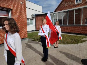 Troje uczniów z pocztu flagowego stoi w rzędzie ustawionym wzdłuż budynku szkoły. Między uczniami zachowany jest odstęp. Wszyscy są ubrani w bialo- czarne stroje i przepasani wstęgami w kolorach polskiej flagi. Uczeń stojący po środku trzyma flagę Polski.