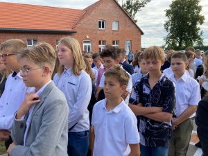 Uczniowie naszej szkoły w strojach galowych stoją w rzędach na szkolnym boisku. W tle widać budynek szkoły.