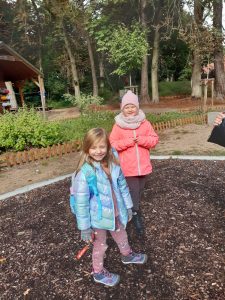 Dwie dziewczynki stoją na placyku wysypanym kamieniami i przykrytym ziemią. W tle widać las.