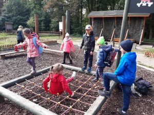 Uczniowie podczas zabawy na placu przy szkole leśnej na Barbarce. Dzieci bawią się przy siatce zamontowanej tuż nad ziemią. Jedna z uczennic siedzi na niej a r5 dzieci stoi w około.