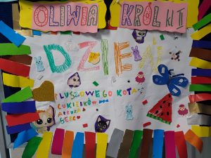 Zdjęcie plakatu kandydatki do małego Samorządu Uczniowskiego. U góry plakatu napis :"OLIWIA KRÓL KL II a pod nim kolorowymi pisakami :" DZIEŃ PLUSZOWEGO KOTA CUKIERKÓW ARBUZA SERC". Plakat ozdobiony kolorwymi rysunkami i paskami przyklejonymi po całym obwodzie.