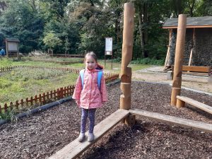 Uczennica stoi na desce umieszczonej tuż nad ziemią. W tle widać drzewa i fragment tężni. Dziewczynka stoi z wyprostowanymi wzdłuż ciała rękami. Na plecach ma plecak.