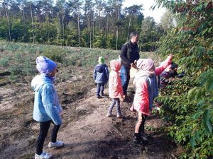 Uczniowie przygladają się drzewom rosnącym w szkólce lesnej na barbarce. Wsród uczniów stoi Pan opowiadający im o rosliności szkółki.