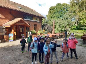 Uczniowie pozują do zdjęcia przed jednym z budynków na terenie Barbarki. Częśc uczniow ma podniesione w górę ręce. Buden stojacy za nimi jest wykonany z cegły o drewna. W tle widać drewniane ławeczki i drzewa.