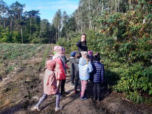 Uczniowie stoją na polanie otoczonej drzewami. Przed nimi stoi mężczyzna wskazujący na wprost siebie palcem.