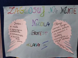 Zdjęcie plakatu kandydatki do małego Samorządu Uczniowskiego. Na niebieskim tle kolorowy napis :"ZAGŁOSUJ NA MNIE NIKOLA ERNEST KLASA III". Po bogach na wycietych z białego papieru szkrzydłach następujące informacje: "Jestem: - miła, - sympatyczna, -koleżeńska, - usmiechnięta, -zawsze służę pomocą" oraz "Oferuję: -pomoc innym, - wspólna zabawa na przerwach i świetlicy, - pomaganie w różnych zbiorkach charytatywnych".