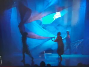 Zdjecie wykonane podczas przedstawienia w Baju Pomorskim. Na rozmytem niebieskim tle widac postaci trojga aktorówtrzymających zawieszoną u góry sceny tkaninę.