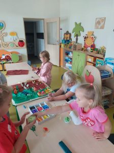 Uczennice siedzą bądź stoją przy połączonych stołach, na których rozłożono kolorowe plasteliny i przybory plastyczne. Miedzy nimi leży kolorowa makieta nad którą pracują uczniowie.