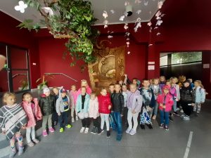 Uczniowie stoją na korytarzu w Baju pomorskim. W tlewidac czerwona ścianę i zwisające z sufityu białe papierowe kwiatki i fragment drzewa.