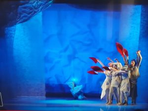 Zdjęcie wykonane podczas przedstawienia w Baju pomorskim. Na niebieskim tle stoją oboksiebie aktorzy trzymający w dłoniach czerwone, podłużne kartki.