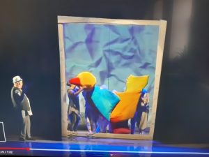 Zdjęcie wyświetlanego filmu. Na ciemnym tle stoi aktor patrzący na brązową framugę w której stoją aktorzy podtrzymujacy kukiełkę ptaka.