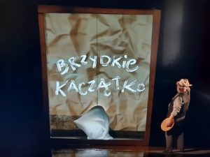 Zdjęcie wykonane podczas przedstawienia W Baju Pomorskim. Na czarnym tle drewniana framuga otaczajaca brązowy lekko pognieciony papier z napisem BRZYDKIE KACZĄTKO. Obok stoi aktori patrzy w stronę napisu.