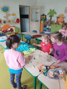 Trzy dziewczynki stoją przy połączonych stolikach w swojej sali lekcyjnej. N stolikach leży kolorowa makieta nad która pracują dziewczynki oraz przybory plastyczne, kolorowe muliny i plastelina.