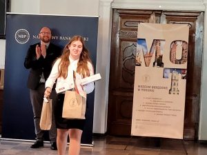 Paulina Ernest idzie z otrzymanami nagrodami. Trzyma dwie papierowy torby i dyplom. WStojący w tle mężczyzna bije jej brawo.