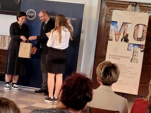 Uczennica Paulina Ernest otrzymuję nagrodę za udział w konkursie. Dziewczyna stoi plecami do zdjęcia a przed nią stoi mężczyzna, który odbiera papierową torbę od stojącej obok kobiety. Obok wisie= plakat okręgowego muzeum w Toruniu.