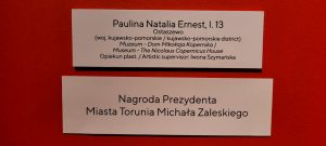 Zdjęcie dwóch tabliczek umieszczonych na czerwonym tle - na pierwszej napis: "Paulina Natalia Ernest, l.13 Ostaszewo (woj. kujawsko-pomorskie/kujawsko-pomorskie district) Muzeum- Dom Mikołaja Kopernika/ Museum - The Nicolaus Copernicus House Opiekun plast. / Artistic supervisor: Iwona Szymańska". Na drugiej tablicze umieszczonej pod spodem napis: "NAGRODA PREZYDENTA MIASTA TORUNIA MICHAŁA ZALEWSKIEGO".