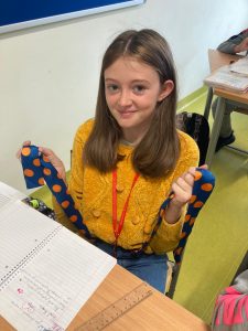 Uczennica siedzi przy stoliku. Usmiecha się. W rękach trzyma granatowa szarfę w żółte kropki.
