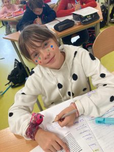 Zdjęcie uczennicy rozwiązującej zadania. Dziewczynka ma na sobie bluzzę ozdobioną czarnymi kropkami a na policzkach naklejone kolorowe kropki imitujące piegi.