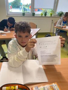 Zdjęcie ucznia siedzącego przy stoliku i trzymajacego w opartych na stole rękach kartę pracy przygotowaną z okazji dnia kropki.