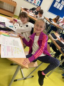 Uczennica siedzi przy stoliku i podnosi w górę nogę aby pokazać getry w białe kropki. W tle widać pracujących uczniów.