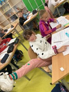 Uczennica siedząca przy szkolnym stoliku wyciąga w górę nogę by pokazać ozdobione kropkami spodnie. W tle widać pracujących uczniów.