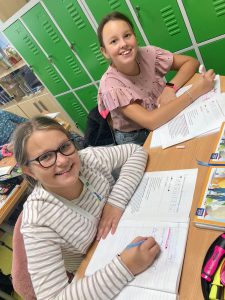 Uczennice uśmiechają się do zdjęcia. Dziewczynki siedzą przy wspólnej ławce i trzymają w dłoniach dlugopisy.