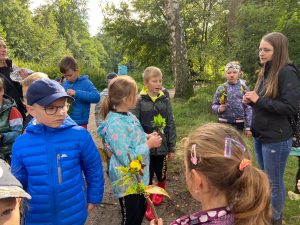 Dzieci stoją na leśnej polanie wokół Pani. Uczniowie trzymają w dloniach liscie.