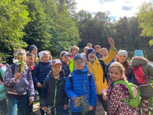 Uczniowie stoją na lesnej polanie. W dłoniach trzymają liście.