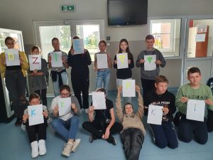 Uczniowie ustawieni na szkolnym korytarzu trzymaja kartki z literami tworzącymi napis DZIEŃ KROPKI.