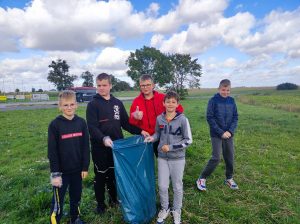 Piątka uczniów klasy siódmej pozuje do zdjęcia na trawiku obok szkolnego orlika. Dwóch chłopców trzyma niebieski worek ze śmieciami. Uczeń stojący za workiem ma podniesiony w górę kciuk przy widocznej lewej dłoni.