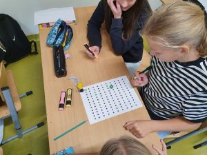 Uczennice klasy szóstej siedzą przy szkolnej ławce, na której leży kartka z ustawionymi w rzędy kropkami. Dziewczyny trzymają w dłoniach kolorowe pisaki.