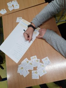 Zdjęcie biurka na którym leżą karteczki z działaniami wykonanymi przez uczniów. Na zdjęciu widać jak jeden z uczniów sprawdza zadania i zapisuje na dużej kopecie dane uczniów, którzy poprawnie je wykonali..