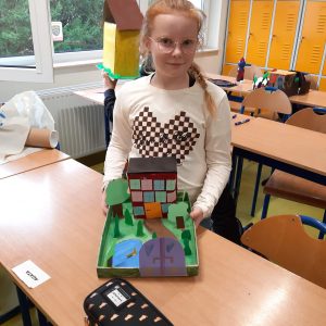 Uczennica klasy czwartej stoi przy ławce i prezentuje wykonaną przez siebie makietę Akademii Pana Kleksa. Makieta przedstawia budynek z wieloma oknami, otoczony drzewami i stawem.