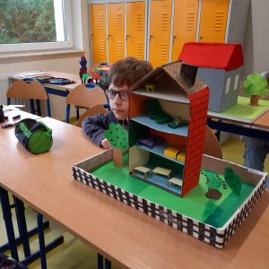 Uczeń klasy czwartej siedzi przy stoliku lekko chowając się za ustawioną na nim makietą Akademii Pana Kleksa. Makieta przedstawia przekrój trzypoziomowego budynku ze spadzistym dachem. na każdym piętrze można dostrzec miniaturowe meble.